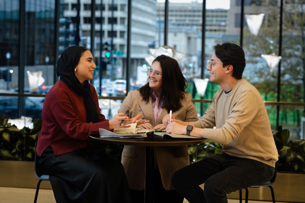 Two young women and a man sit at a table with pens and hand while talking