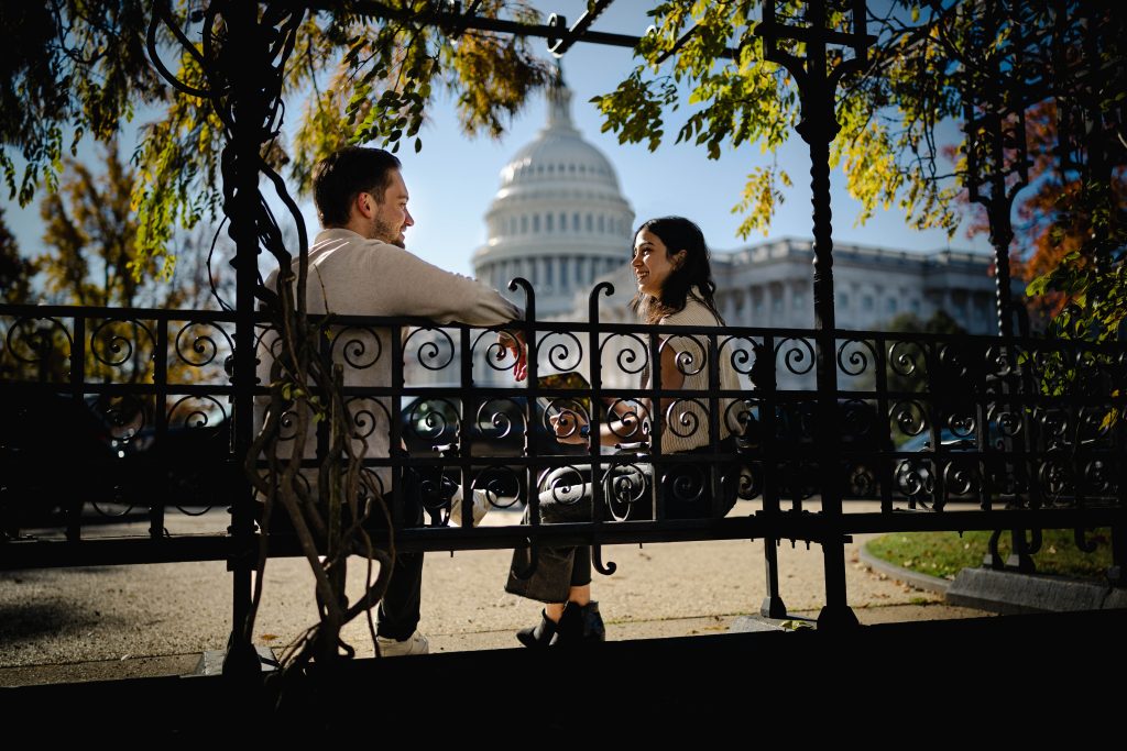 Young man and woman sit at a bench with the US Capitol in the background on a clear sunny day