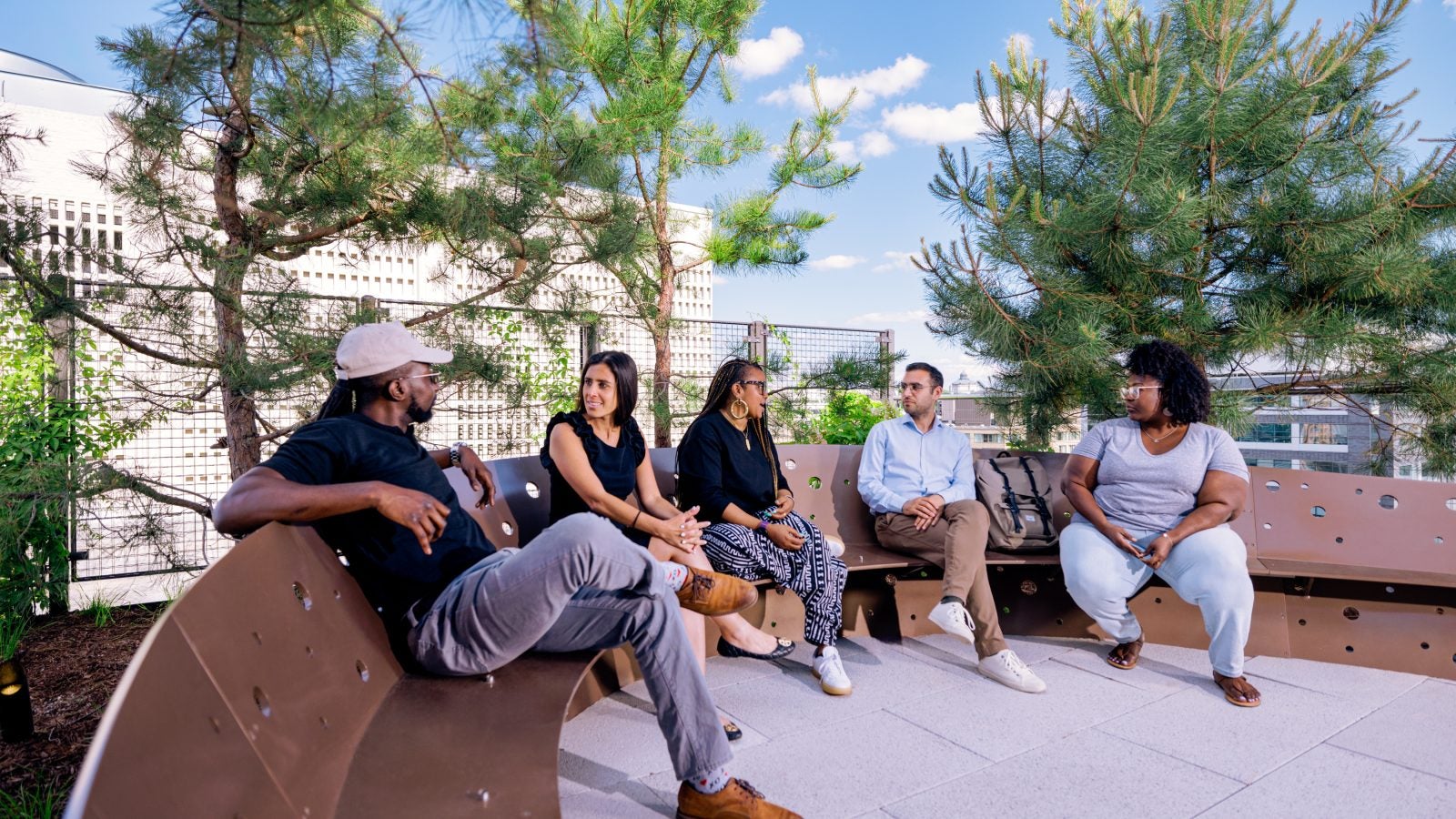 Five students hanging out and chatting on a curved, metal bench on a rooftop. Green trees surround them.