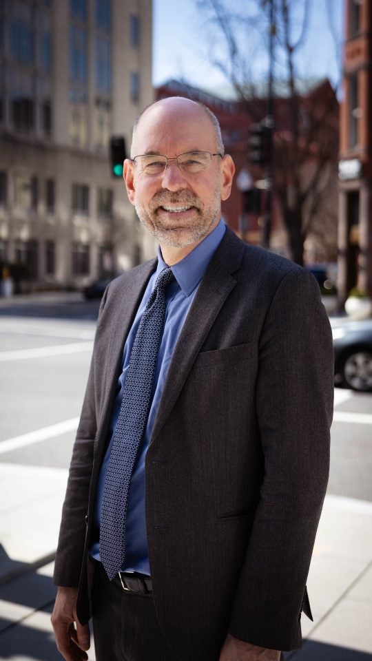 shot of Uwe, who wears a suit and tie, and is standing on a city corner
