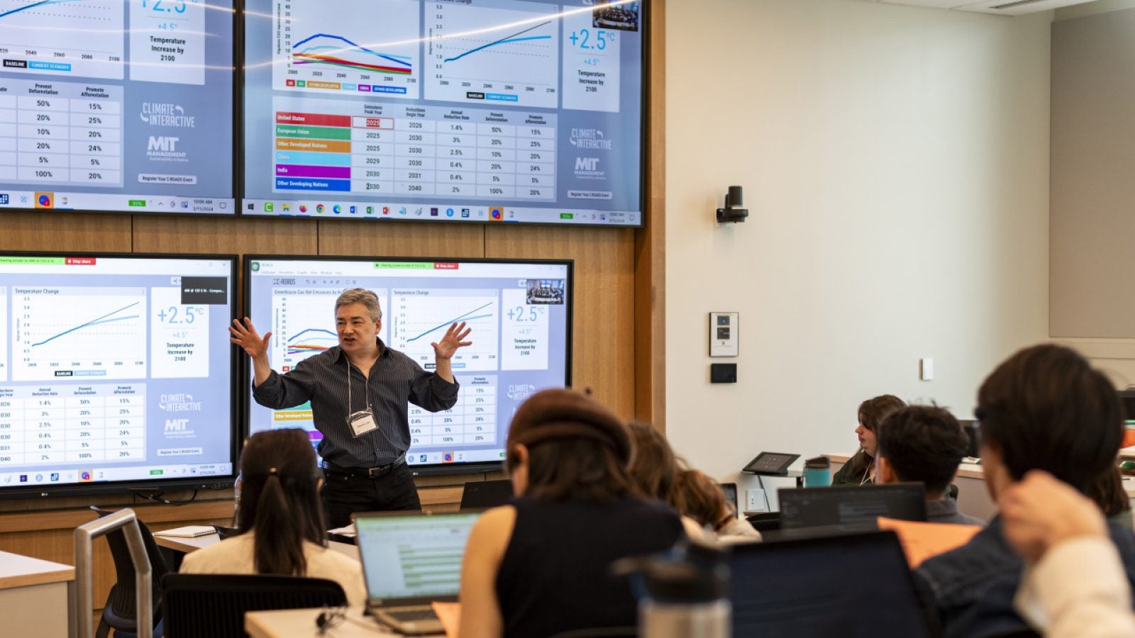 A professor talking a to a group of students in a classroom with four screens behind her.