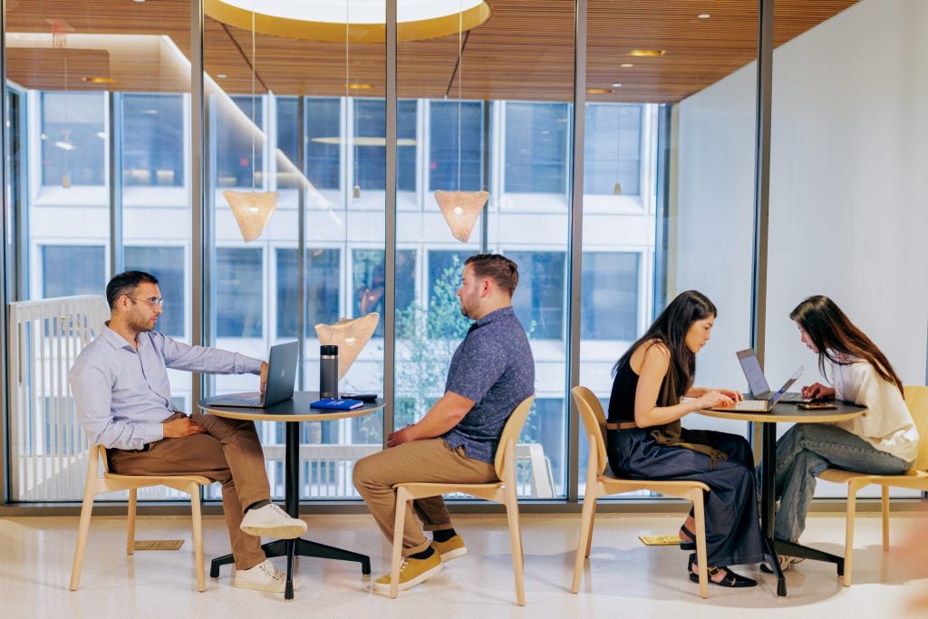 Two people sitting and talking over a small table indoors with two young women in the background working.